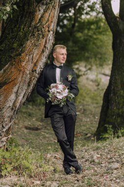 Takım elbiseli ve kravatlı bir adam bir ağacın yanında elinde bir buket çiçek tutuyor. Sahne sakin ve huzur dolu. Uzağa bakan adamla birlikte.