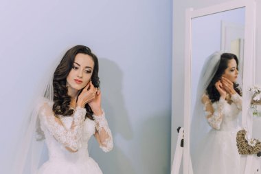 Bir kadın düğünü için hazırlanıyor ve aynada kendine bakıyor. Beyaz bir elbise ve duvak takıyor.