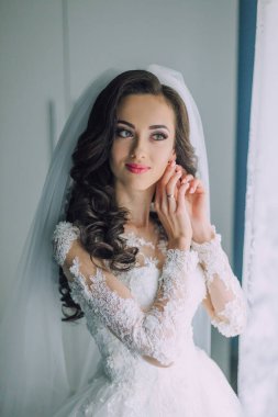 Gelinlik giymiş bir kadın kameraya bakıyor. Peçe takıyor ve elleri kulaklarında.