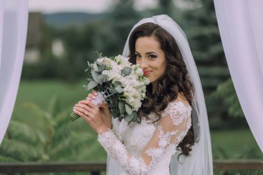 Bir gelin bir buket çiçek tutuyor ve gülümsüyor. Gelin beyaz bir gelinlik ve duvak takıyor. Çiçekler beyaz ve yeşil ve gelin beyaz bir perdenin önünde duruyor.