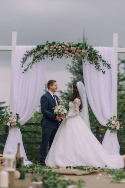 Gelin ve damat beyaz kemer yolunun altında durur, gelin beyaz elbise giyer ve damat mavi takım giyer.