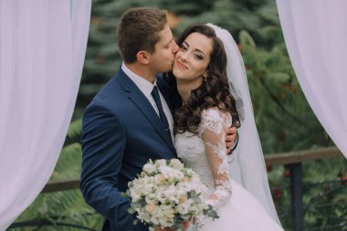 Bir gelin ve damat beyaz bir perdenin önünde birbirlerinin dudaklarını öpüyor. Gelin beyaz bir elbise giyiyor ve damat mavi bir takım giyiyor.