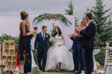 Bir gelin ve damat nikah masasında yürüyorlar. Gelin beyaz bir elbise giyiyor ve damat mavi bir takım elbise giyiyor. Misafirler yeni evliler için alkış ve tezahürat yapıyorlar.