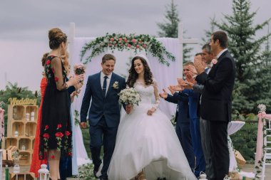 Bir gelin ve damat nikah masasında yürüyorlar. Gelin beyaz bir elbise giyiyor ve damat mavi bir takım elbise giyiyor. Misafirler yeni evliler için alkış ve tezahürat yapıyorlar.