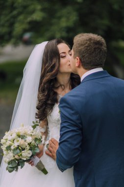 Gelin ve damat beyaz bir duvağın önünde öpüşürler. Gelin elinde bir buket çiçek tutuyor.