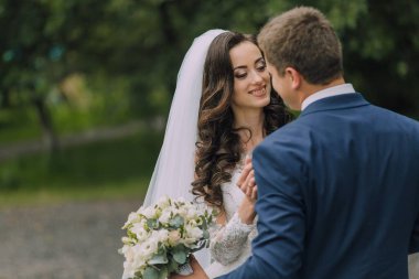 Gelin ve damat parkta duruyorlar, gelin bir buket çiçek tutuyor. Adam mavi bir takım elbise giyiyor ve kadın beyaz bir elbise giyiyor. İkisi de gülümsüyor.