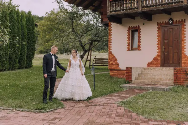 Bir gelin ve damat evin önündeki tuğla yolda yürüyorlar. Gelin beyaz bir elbise giyiyor ve damat siyah bir takım elbise giyiyor. Sahne romantik ve mutlu.