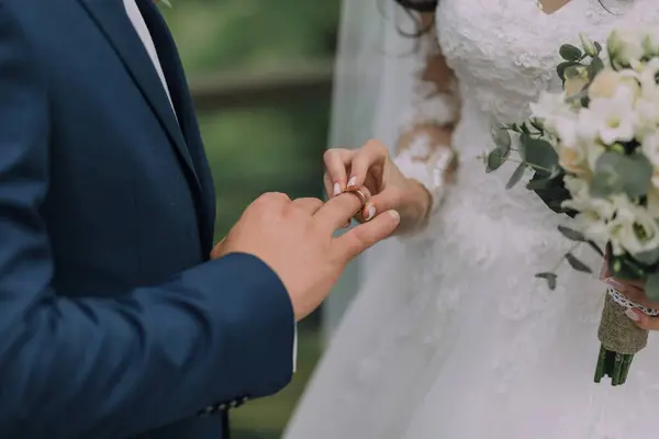 Bir erkek ve bir kadın evleniyor ve erkek kadına bir yüzük vermek üzere. Gelin beyaz bir elbise giyiyor ve damat mavi bir takım elbise giyiyor. Gelin elinde bir buket çiçek tutuyor.