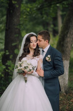 Bir gelin ve damat ormanda sarılıyor. Gelin elinde bir buket çiçek tutuyor.