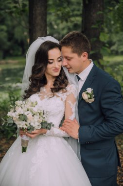 Bir gelin ve damat ormanda fotoğraf çektirmek için poz veriyorlar. Gelin bir buket çiçek tutuyor ve damat kravat takıyor. Sahne romantik ve samimi.