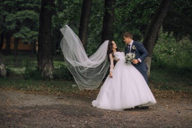 Gelin ve damat bir ormanda duruyorlar, gelin peçe tutuyor ve damat bir buket tutuyor.