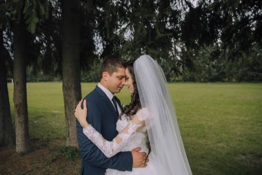 Bir gelin ve damat arka planda ağaçlar olan bir tarlada kucaklaşıyorlar. Sahne romantik ve samimi.
