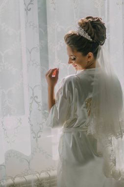 Bir gelin pencerenin önünde duruyor, beyaz bir elbise ve taç giyiyor. Pencereden dışarı bakıyor, muhtemelen manzaraya hayran ya da birini bekliyor.