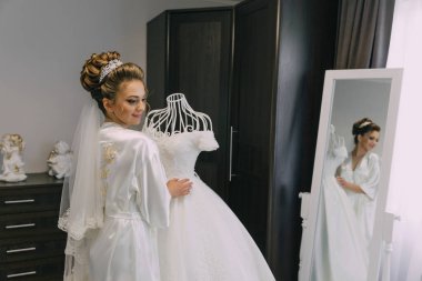 Beyaz elbiseli bir kadın, gelinlik giymiş bir mankenin önünde duruyor. Elbiseyi kollarında tutuyor ve gülümsüyor. Mutluluk ve heyecan kavramı