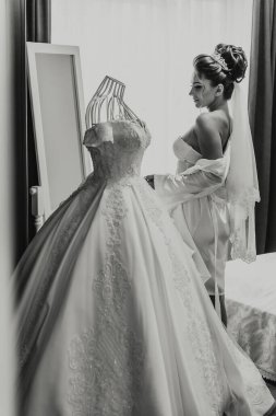 Bir kadın düğüne hazırlanıyor ve üstünde gelinlik olan bir şifoniyerin önünde duruyor.