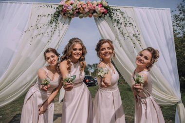 Dört kadın beyaz bir kemerin önünde poz veriyor. Ellerinde çiçekler tutuyorlar ve gülümsüyorlar. Sahne neşeli ve kutlama dolu.