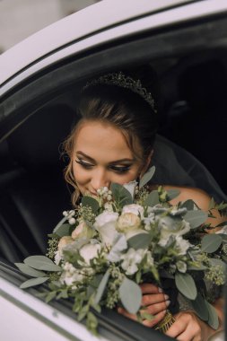 Bir gelin kucağında bir buket çiçekle arabada oturuyor. Gülümsüyor ve mutlu.