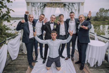 Bir grup adam rıhtımda fotoğraf çektirmek için poz veriyor. Bir adam diz çöküyor ve diğerleri ayakta. Sahne neşeli ve eğlenceli, çünkü erkekler birlikte poz veriyorlar.