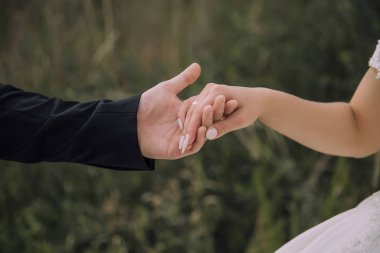 Tarlada el ele tutuşan bir çift. Adam siyah bir takım giyiyor ve kadın beyaz bir elbise giyiyor.