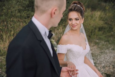 Gelin ve damat tarlada duruyorlar, gelin beyaz elbise giyiyor ve damat siyah takım elbise giyiyor.