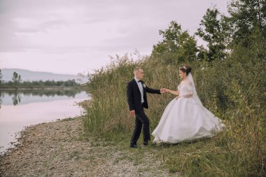 Bir gelin ve damat bir deniz kıyısında yürüyorlar. Gelin beyaz bir elbise giyiyor ve damat siyah bir takım elbise giyiyor. Sahne romantik ve resimli.