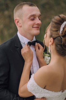 Bir adam bir kadının gelinliğini giymesine yardım ediyor. Adam siyah bir takım elbise ve kravat takıyor. Kadın beyaz bir elbise giyiyor ve saçları topuz şeklinde. Sahne mutlu ve kutlama dolu.