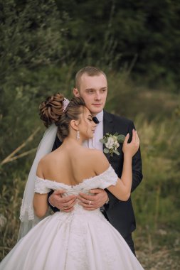 Bir gelin ve damat tarlada kucaklaşıyor. Gelin beyaz bir elbise giyiyor ve damat da takım elbise giyiyor.