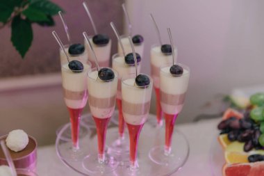 Çeşitli tatlı ve meyvelerle dolu bir masa. İçinde pembe ve beyaz tatlı dolu küçük bardaklar da var.