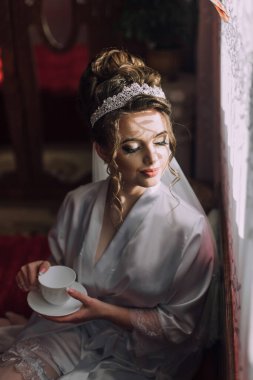Gelinlik giymiş bir kadın elinde beyaz bir kupa tutuyor. Penceresi ve saksısı olan bir odada oturuyor.