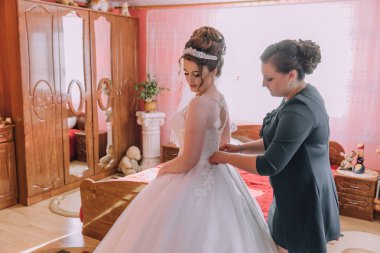 Bir kadın düğünü için hazırlanıyor ve annesi ona yardım ediyor. Gelin beyaz bir elbise giyiyor ve oda pembe bir renk düzeniyle süslenmiş.