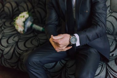 Takım elbiseli bir adam kanepede oturuyor ve saatini ayarlıyor. Kanepe siyah ve üzerinde bir desen var. Adam kravat takıyor ve elinde bir buket çiçek tutuyor.