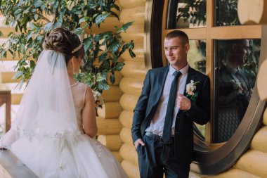 Bir adam ve bir kadın pencerenin önünde duruyorlar. Adam takım elbise ve kravat takıyor ve kadın beyaz bir gelinlik giyiyor. İkisi de gülümsüyor ve birbirlerine bakıyorlar.