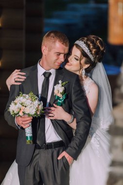 Bir erkek ve bir kadın birbirlerine sarılıyorlar, bir buket çiçek tutan adamla. Kadın gelinlik ve taç takıyor. Sahne mutlu ve romantik.