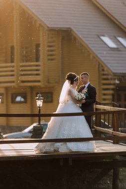 Bir gelin ve damat köprüde el ele tutuşup gülümsüyor. Damat takım elbise giyerken gelin beyaz bir elbise ve peçe takıyor. Sahne sarı bir evin arka planında geçiyor.