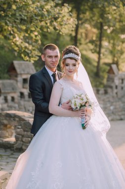 Bir gelin ve damat taş bir duvarın önünde poz veriyorlar. Gelin beyaz bir elbise giyiyor ve elinde bir buket çiçek tutuyor. Adam takım elbise ve kravat takıyor.