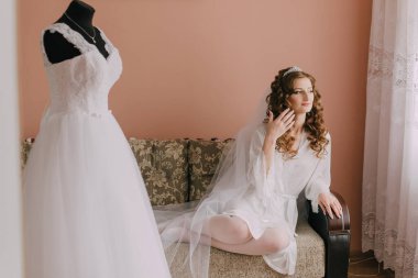 Bir kadın kanepede gelinliğin yanında oturuyor. Duvak ve taç takıyor.