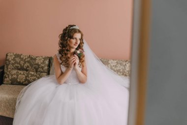 Bir gelin kucağında bir buket çiçekle bir kanepede oturuyor. Beyaz bir elbise ve taç giyiyor.