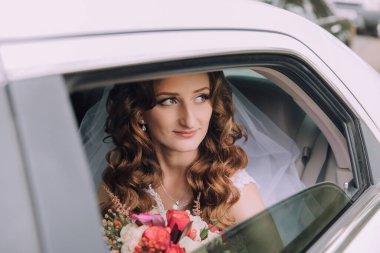 Bir gelin kucağında bir buket çiçekle arabada oturuyor. Gülümsüyor ve pencereden dışarı bakıyor.