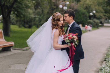 Bir gelin ve damat kaldırımda birbirlerine sarılıyorlar. Gelin beyaz bir elbise giyiyor ve damat bir takım elbise giyiyor. Bir buket çiçek ve çiçek tutuyorlar.