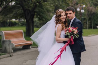 Bir gelin ve damat parkta fotoğraf için poz veriyorlar. Gelin beyaz bir elbise giyiyor ve damat bir takım elbise giyiyor. Bir buket çiçek ve çiçek tutuyorlar.