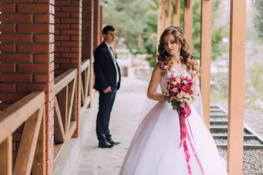 Bir gelin ve damat tuğla bir duvarın önünde duruyor. Gelin elinde bir buket çiçek tutuyor.