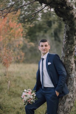 Takım elbise giymiş ve kravat takmış bir adam elinde bir buket çiçekle bir ağacın yanında duruyor. Bir fotoğraf için poz veriyor, muhtemelen bir düğün ya da diğer özel durumlar için.