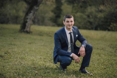 Takım elbiseli ve kravatlı bir adam çimlerin üzerinde oturuyor. Gülümsüyor ve kameraya bakıyor.