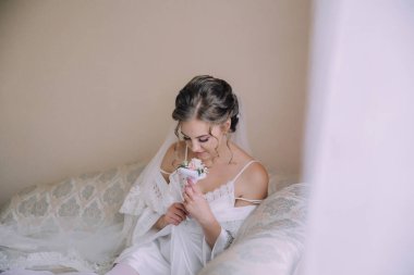 Bir kadın beyaz bir battaniyeyle bir yatakta oturuyor ve elinde bir çiçek tutuyor. İmajın yumuşak ve romantik bir havası var. Kadının zarif kıyafetleri ve elinde narin bir çiçek var.