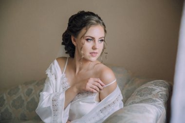 Beyaz elbiseli bir kadın kanepede oturuyor. Dantelli bir bluz giyiyor ve kolunu vücuduna dolamış. İmajın romantik ve zarif bir havası var.