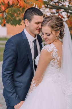 Bir gelin ve damat fotoğraf için poz veriyor. Gelin beyaz bir elbise giyiyor ve damat mavi bir takım elbise giyiyor. İkisi de gülümsüyor ve kameraya bakıyor. Sahne mutlu ve romantik.