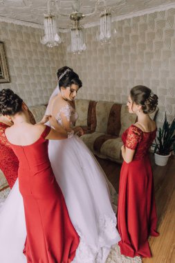 Üç kadın düğün için hazırlanıyor. Biri beyaz elbise giyiyor ve diğer ikisi kırmızı elbise giyiyor.