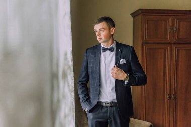 Takım elbiseli ve kravatlı bir adam pencerenin önünde duruyor. Papyon ve cep mendili takıyor.