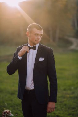 Takım elbiseli ve kravatlı bir adam tarlada duruyor. Kravatını düzeltiyor ve kameraya bakıyor. Adam takım elbise ve kravat giydiği için resmiyet ve zarafet kavramı.