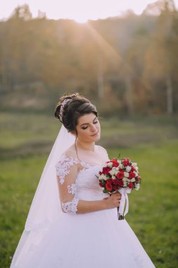 Bir gelin elinde bir buket kırmızı çiçekle bir tarlada duruyor. Sahne sakin ve huzurlu, gelin kameraya bakıyor.